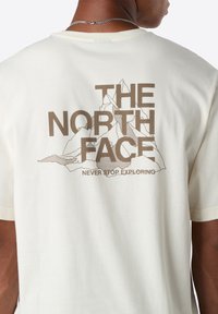 Personne portant un T-shirt blanc avec un logo marron "THE NORTH FACE" et un graphisme de montagne, incluant la phrase "NEVER STOP EXPLORING" au dos.