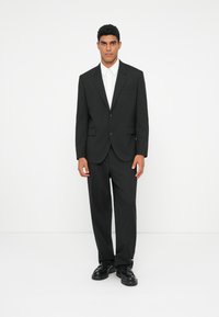Traje negro con chaqueta de un solo botón, solapas con muesca y pantalones a juego; llevado sobre una camisa blanca de botones y zapatos negros.
