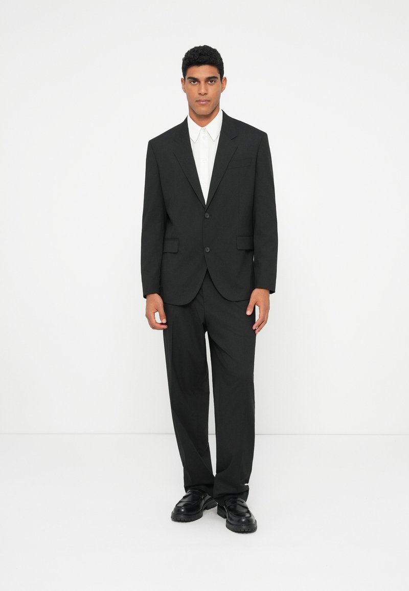 Traje negro con chaqueta de un solo botón, solapas con muesca y pantalones a juego; llevado sobre una camisa blanca de botones y zapatos negros.