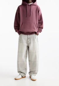 Persona che indossa una felpa oversize color rosa polveroso, jeans larghi grigio chiaro con toppe e sneakers bianche con strisce nere e suola in gomma.
