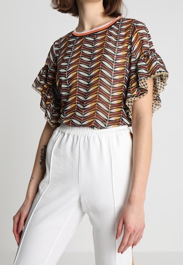 Femme portant un chemisier à motifs marron, orange et blanc avec des manches à volants, ainsi qu'un pantalon blanc taille haute avec une ceinture élastique.
