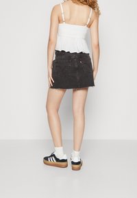 Žena v bílém bezrukávovém topu, černé denimové minisukni, bílých ponožkách a černo-bílých teniskách, stojící před jednoduchým pozadím.