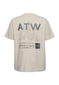 Beige bomulds t-shirt med "ATW" i store grå bogstaver og et bjerggrafik på ryggen, med forskellige tekstelementer og logoer.