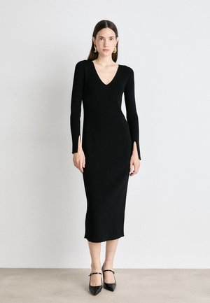 Black ribbed knit dress with a v-neckline, long sleeves, and side slits. Worn with black heels featuring ankle straps.