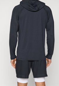 Schwarzer langärmeliger Sport-Hoodie mit Kapuze, glatter Textur und Raglanärmeln. Kombiniert mit dunklen Shorts mit perforierten Akzenten.