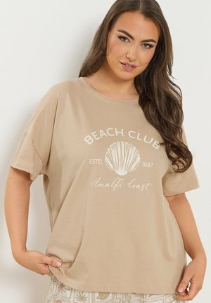 Vrouw met lang bruin haar, gekleed in een beige T-shirt met opschrift "Beach Club Amalfi Coast", houdt de zoom met beide handen vast tegen een effen achtergrond.