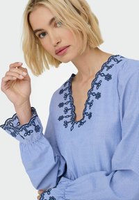 Blouse rayée bleu clair avec des broderies florales bleu marine le long de l'encolure et des poignets, avec des manches amples et flottantes et un décolleté en V.