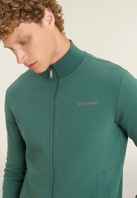 Giacca verde con zip realizzata in tessuto liscio; presenta un colletto alto, tasche laterali e una zip argentata con un logo sul petto.