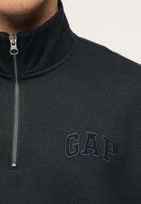 Κοντινό πλάνο ενός ατόμου που φοράει μαύρο φούτερ με φερμουάρ και ασημί φερμουάρ, με ανάγλυφο λογότυπο «GAP» στο στήθος.