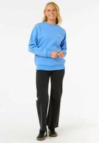 Sudadera azul clara con puños y dobladillo de canalé, que presenta un pequeño logotipo. Combinada con pantalones negros y zapatillas negras con suelas marrones.
