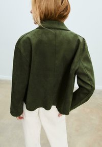 Giacca in finta suede verde con texture liscia, colletto a risvolto, spalle cadute e orlo scallopato, indossata sopra pantaloni bianchi.