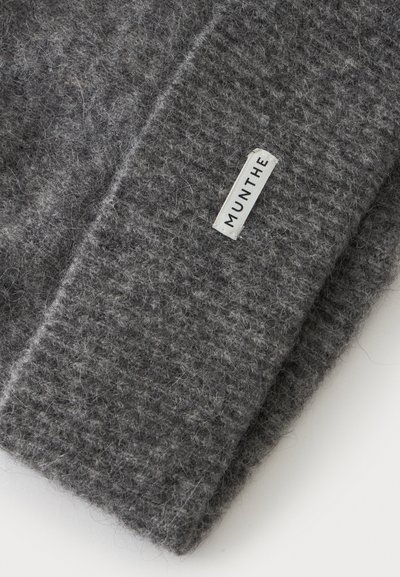 Gros plan d’un tissu tricoté gris texturé avec une étiquette rectangulaire blanche indiquant "MUNTHE" cousue sur le matériau.