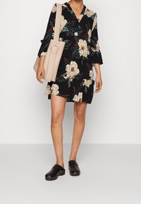 Abito nero a fiori con maniche lunghe, caratterizzato da grandi fiori color crema e foglie verdi. Abbinato a una borsa beige e a zoccoli neri.