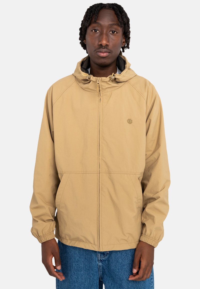 Beige polyesterjakke med hætte, frontlynlås, elastiske manchetter og sidelommer. Har et lille logo på venstre brystområde.