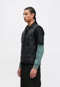 Chaleco acolchado negro con cierre de cremallera, usado sobre una camisa oscura y una camiseta de manga larga verde. Diseño sencillo y textura moderada.