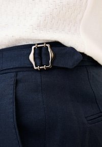 Nahaufnahme eines marineblauen Stoffgürtels mit silberner rechteckiger Schnalle, getragen über passenden Hosen und einem weißen, strukturierten Strickoberteil.