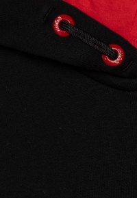 Sweatshirt à capuche avec un corps noir, une accent rouge en haut, un cordon noir passant par des œillets rouges, et un tissu texturé.