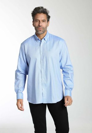 Homme aux cheveux poivre et sel portant une chemise bleu clair boutonnée et un pantalon noir, debout devant un fond clair uni.