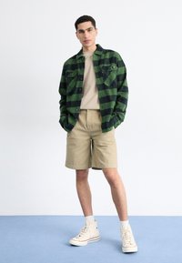 Grøn og sort ternet flannel skjorte, beige t-shirt, khaki shorts og cremefarvede hæl-sneakers. Står på en blå tæppebelagt overflade.