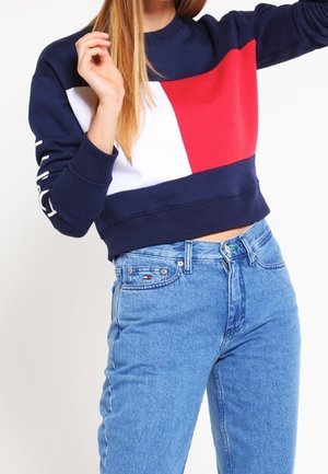 Mujer con sudadera de manga corta en bloques de color azul marino, rojo y blanco, combinada con vaqueros de talle alto azules con logo de la marca en el bolsillo.