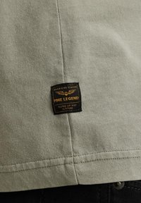 Camiseta de algodón verde claro con una etiqueta cuadrada negra con texto dorado y alas, cosida en el dobladillo inferior.