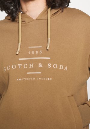 Personne portant un sweat à capuche marron avec des cordons beige et un texte indiquant "1985 Scotch & Soda Amsterdam Couture" sur le devant.