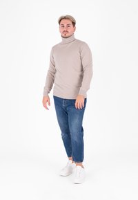 Maglione beige con collo alto e polsini e orlo a costine, abbinato a jeans slim fit blu e sneakers bianche. Il modello è in piedi contro uno sfondo bianco.