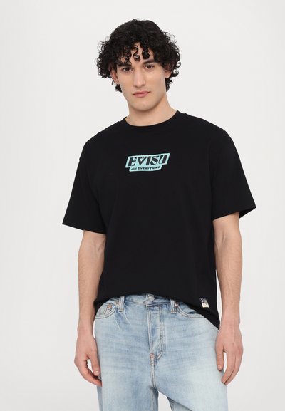 Melna kokvilnas T-krekls ar īsām piedurknēm, ar turkīza grafisko logotipa tekstu "EVISU izdarīja VISS" priekšpusē. Stilizēts ar gaiši ziliem džinsiem.