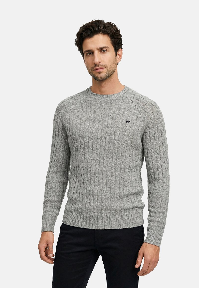 Mann trägt einen hellgrauen, kabelgestrickten Pullover mit einem kleinen gestickten Logo auf der Brust und schwarze Hosen, steht vor einem schlichten Hintergrund.