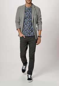 Grijze blazer over een geometrisch patroon van een blauwe en zwarte t-shirt, gecombineerd met donkere broek en zwarte sneakers met witte veters.