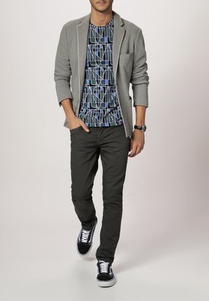 Blazer grigio sopra una t-shirt blu e nera con motivo geometrico, abbinato a pantaloni scuri e sneakers nere con lacci bianchi.