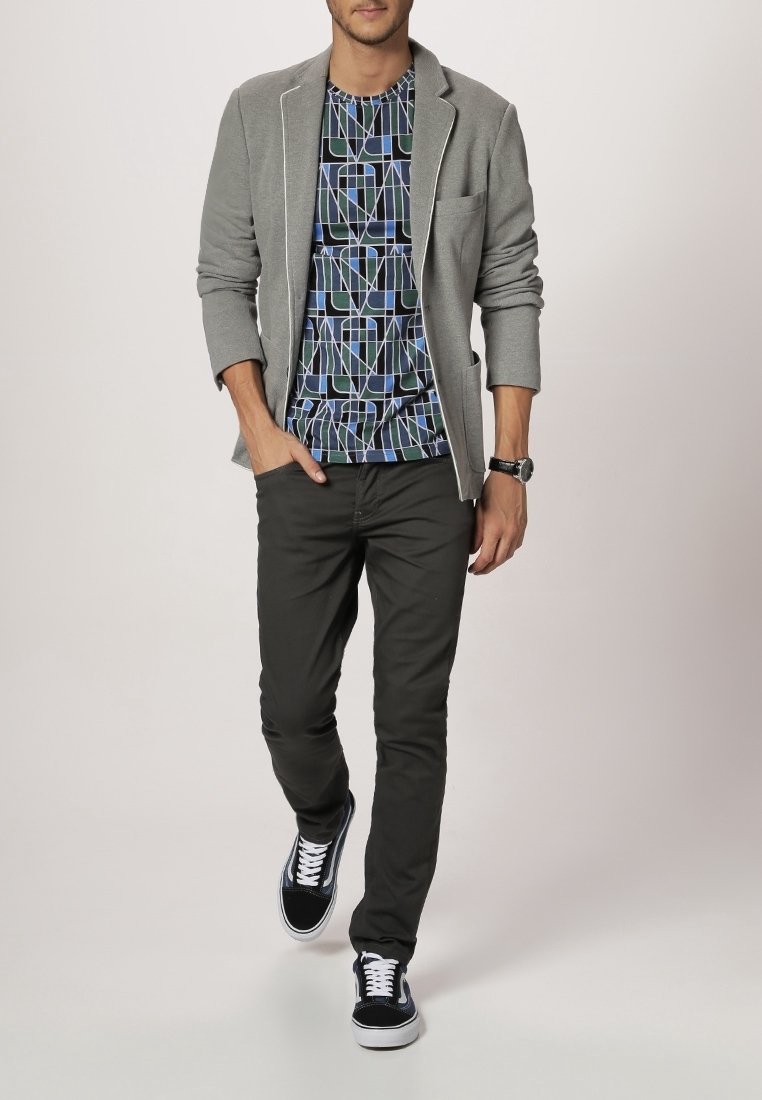 Grijze blazer over een geometrisch patroon van een blauwe en zwarte t-shirt, gecombineerd met donkere broek en zwarte sneakers met witte veters.