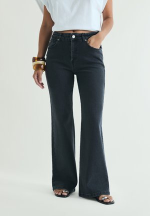 Woman wearing high-waist black wide-leg jeans, white cropped top, two chunky bracelets, and black open-toe sandals with silver rings.