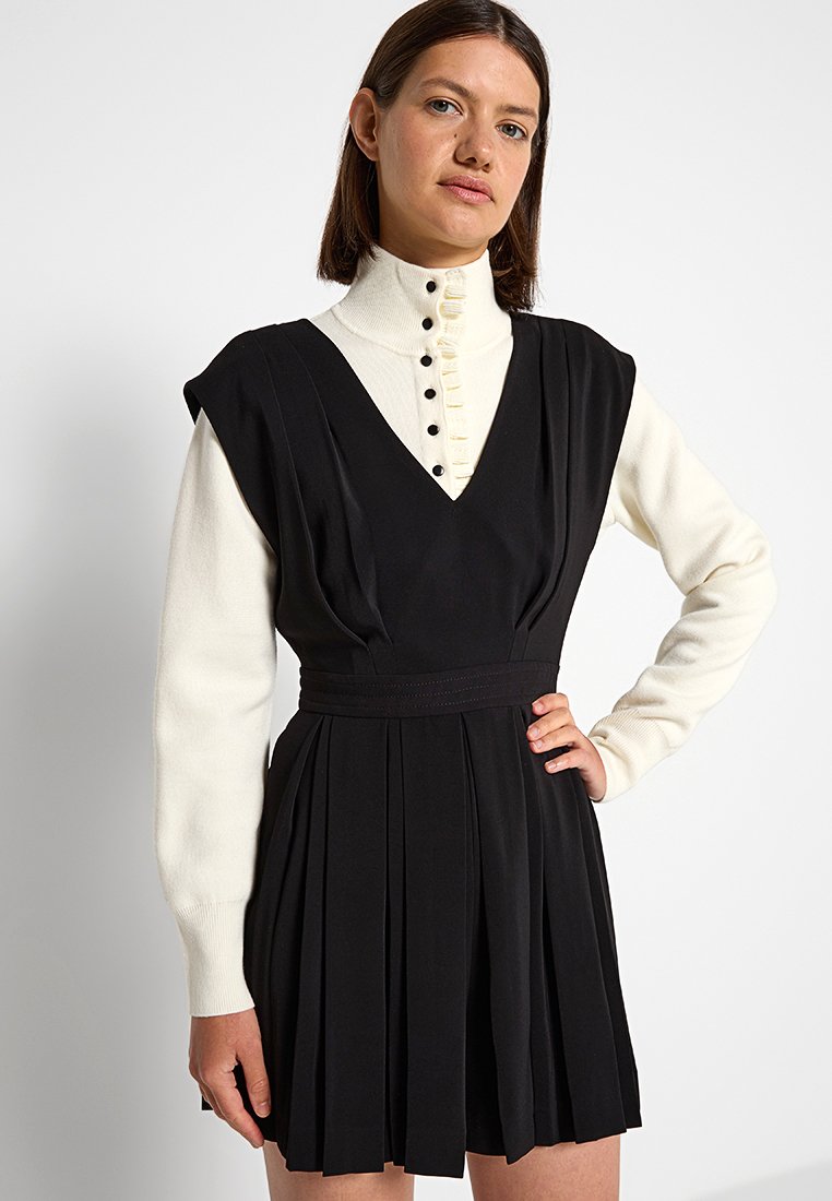 Schwarzes plissiertes Kleid mit tiefem V-Ausschnitt, das eine taillierte Taille aufweist und von einem kontrastierenden weißen Hochkragenoberteil mit langen Ärmeln und Knopfakzenten ergänzt wird.