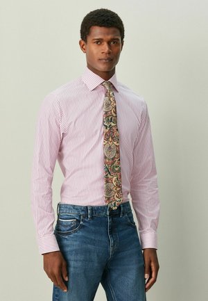 Gestreiftes rosa Hemd mit Druckknopfkragen, kombiniert mit einer bunt gemusterten Paisley-Krawatte. Dazu blaue Jeans getragen.