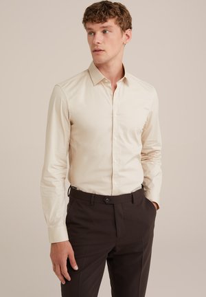 Camisa de botones beige claro con cuello clásico y mangas largas, combinada con pantalones marrón oscuro. Tejido suave, ajuste entallado.