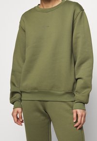 Olivgrön sweatshirt med rund halsringning, ribbade muddar och nederkant. Har en liten logotyp centrerad på bröstet. Mjuk, texturerad tyg.
