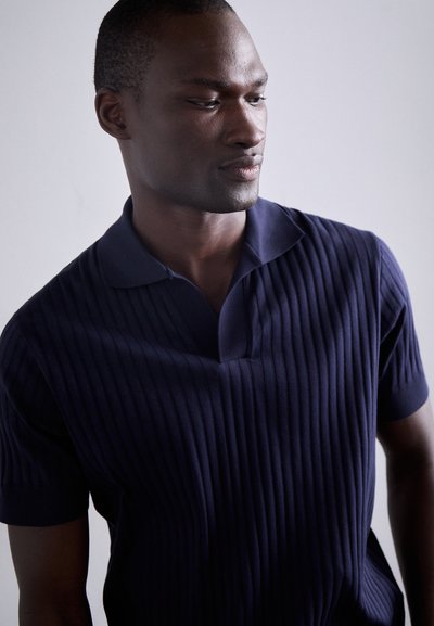 Polo-shirt côtelé bleu marine avec un col court et un décolleté en V prononcé. Le tissu a une texture lisse avec des rayures verticales.