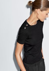 Zwarte t-shirt met korte mouwen van gladde stof, met een ronde halslijn en decoratieve zilveren knopen op de schouder, gecombineerd met donkere broek.