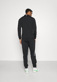 Survêtement de sport noir comprenant une veste zippée et un pantalon fuselé. Rayures blanches contrastantes sur les côtés. Tissu doux et lisse.