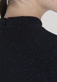 Schwarzes, hochgeschlossenes Kleidungsstück mit einer strukturierten Oberfläche und blauen Sprenklern. Mit einem kleinen schwarzen Knopf im Nacken gesichert.