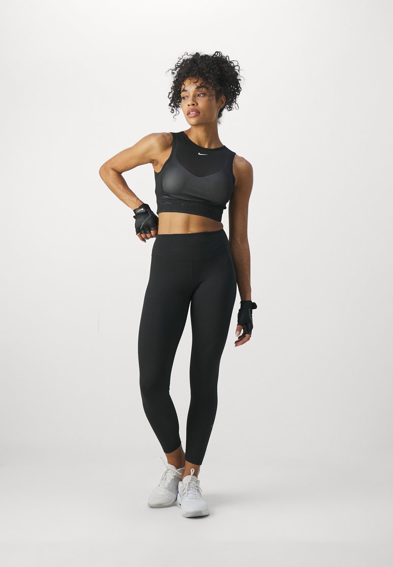 Soutien-gorge de sport noir au design épuré avec logo, associé à un legging noir taille haute. Le modèle porte des gants sans doigts et des baskets blanches.