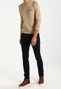 Maglione beige a maniche lunghe con taschino sul petto e cerniera, abbinato a jeans aderenti scuri e sneakers alte marroni.