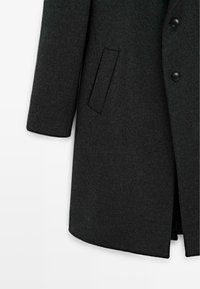 Sezione di un cappotto di lana grigio scuro che mostra una manica lunga, una tasca laterale e due bottoni neri sul pannello frontale.