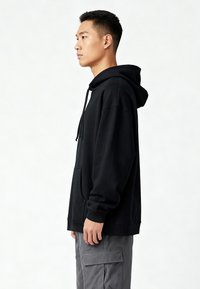 Felpa nera con cappuccio, vestibilità oversize, polsini e orlo a coste, tasca frontale tipo kangaroo, indossata con pantaloni cargo grigi.