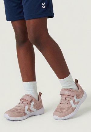 Kind trägt rosa Mesh-Sneakers mit weißen Chevron-Logos, weißen Socken und marineblauen Shorts und steht vor einem schlichten hellen Hintergrund.