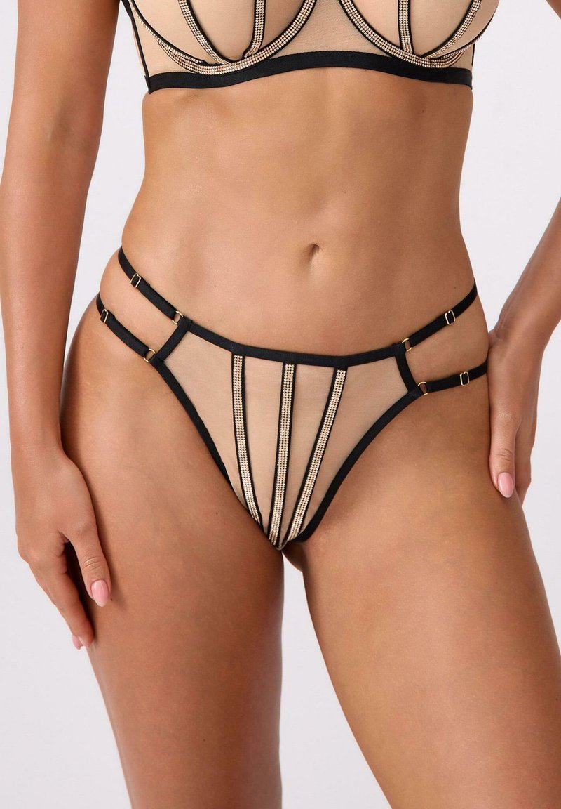 Kobieta ubrana w beżowy i czarny komplet bielizny z przezroczystej tkaniny i pionowymi paskami z kryształków, z podwójnymi ramiączkami na biodrach.