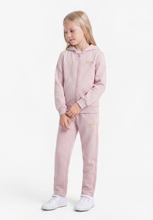 Jeune fille aux longs cheveux blonds portant un survêtement rose Puma et des baskets blanches, debout les mains jointes et regardant vers le bas.