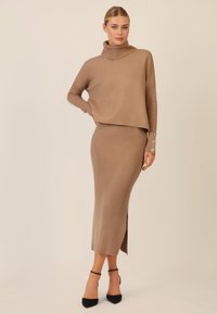 Beige Rippstrickoberteil mit hohem Kragen und enganliegenden Ärmeln, kombiniert mit einem passenden Rippstrick-Midirock mit seitlichem Schlitz, getragen mit schwarzen Absätzen.