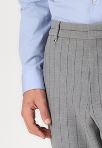 Pantalones de rayas grises hechos de un tejido texturizado, con un frente plano y un lazo para cinturón con un pequeño acento decorativo. Camisa azul clara visible.